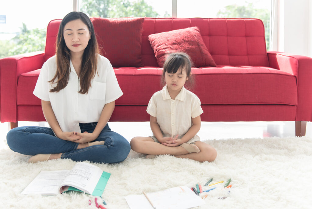 Young Asian mother and little child daughter doing meditation sit on the floor at home, mom teaching cute kid girl learning practice meditation together